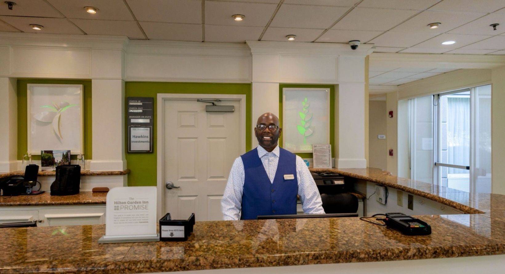 A smiling hotel front desk clerk in a blue vest at the check-in counter