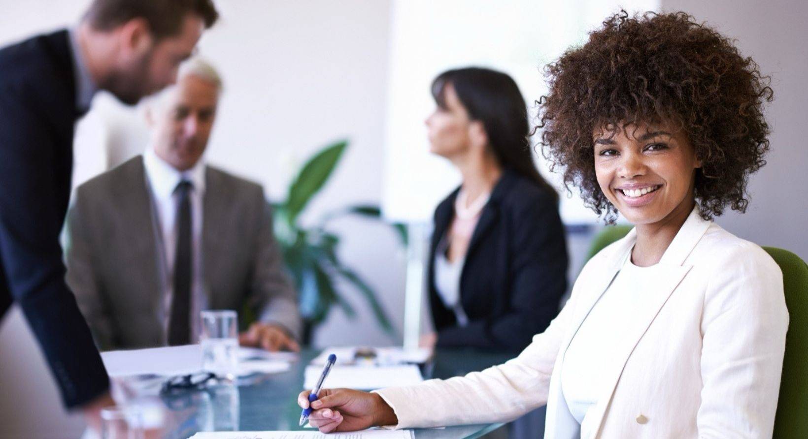 A smiling young professional in a white blazer taking notes during a business meeting
